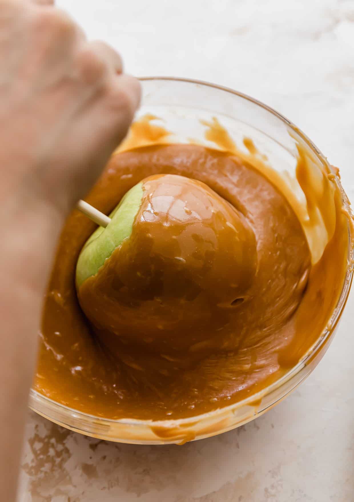 A hand twisting a green Granny Smith apple in a bowl of caramel.