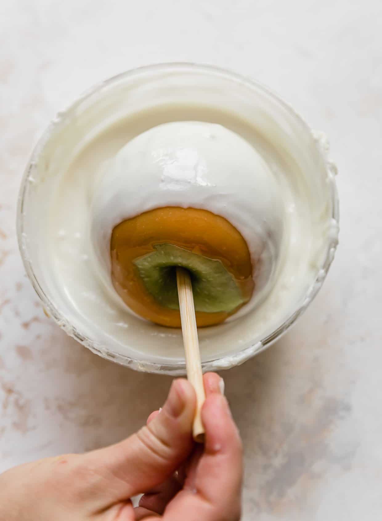 A caramel coated apple being dipped in a bowl filled with melted almond bark (or white chocolate).