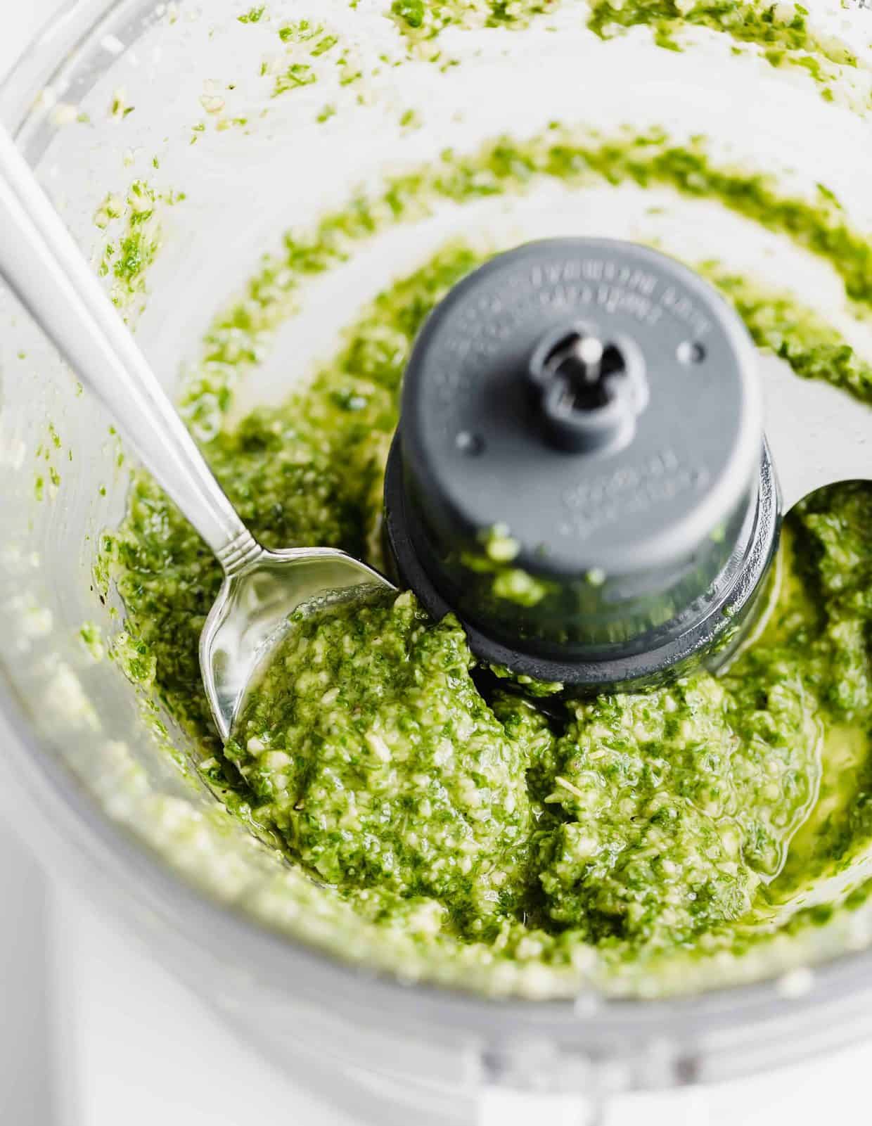 A spoon scooping up fresh basil pesto from a food processor.