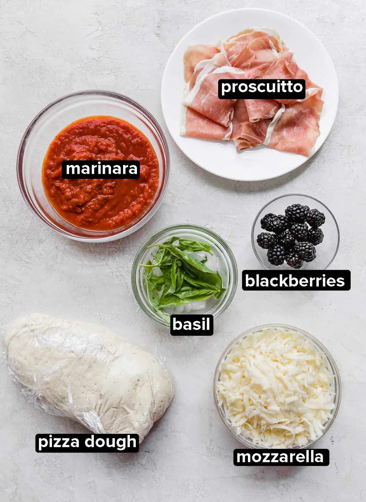 Blackberry Ricotta Pizza ingredients on a gray background: pizza dough, cheese, basil, blackberries, prosciutto, marinara.