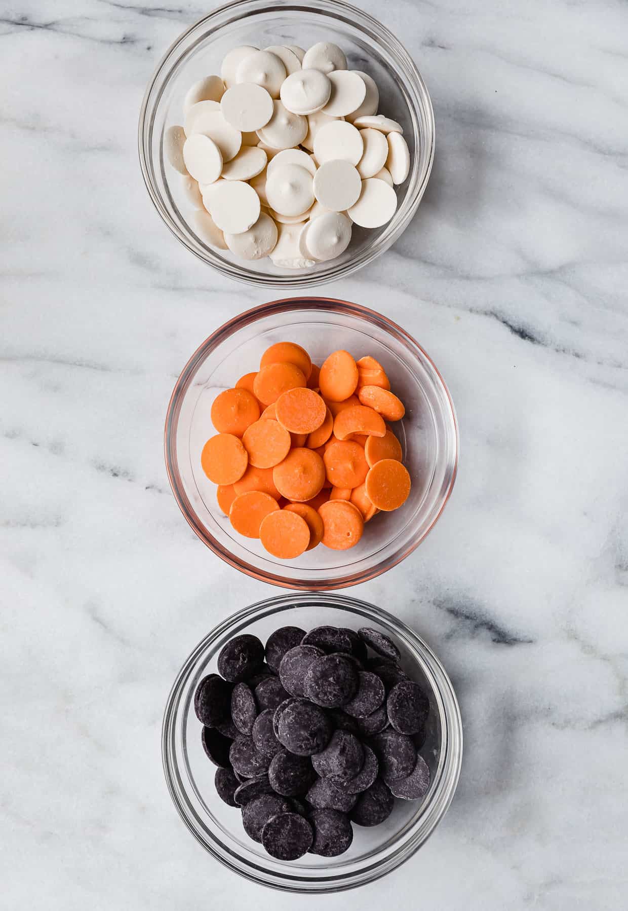 Three glass bowls filled with black, orange, and white melting chocolate disks on a white marble table.