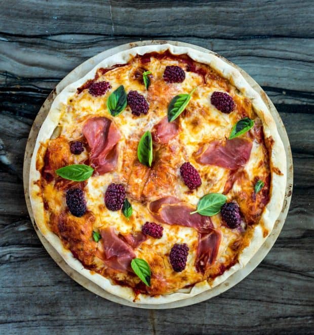 A blackberry and prosciutto topped pizza on a grey background.