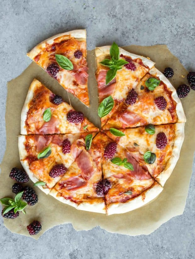 A pizza garnished with fresh basil, prosciutto, and fresh blackberries. 