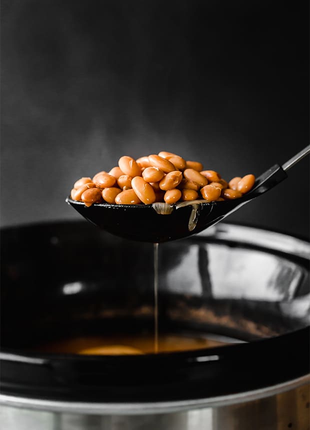 A slotted spoon lifting up a spoonful of cooked pinto beans from the slow cooker, with some liquid dripping through the spoon.