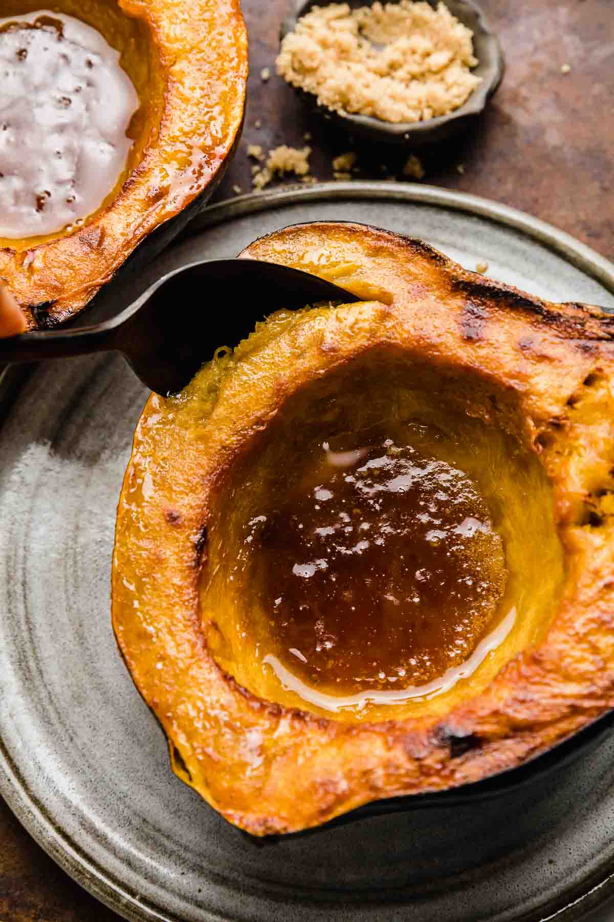 Roasted acorn squash with buttery brown sugar in the center of the squash, and a spoon scooping out the squash flesh.