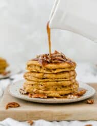 A stack of pumpkin pancakes with chopped pecans on top and syrup being drizzled over the stack.