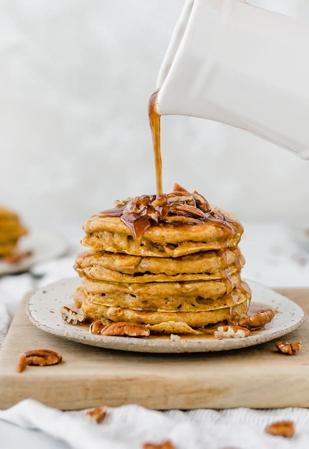 A stack of pumpkin pancakes with chopped pecans on top and syrup being drizzled over the stack.