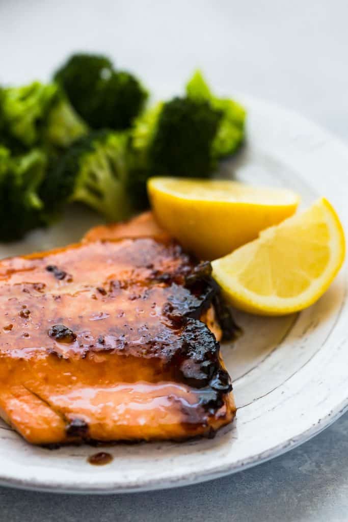 salmon fillet on a white plate, with a brown sugar crusted top. Lemons and broccoli in the background. 
