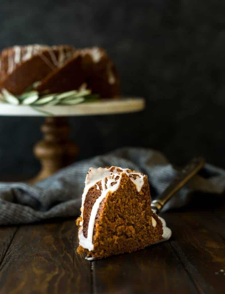 A slice of gingerbread bundt cake in the foreground and the remainder of the cake on a cake stand in the background. 