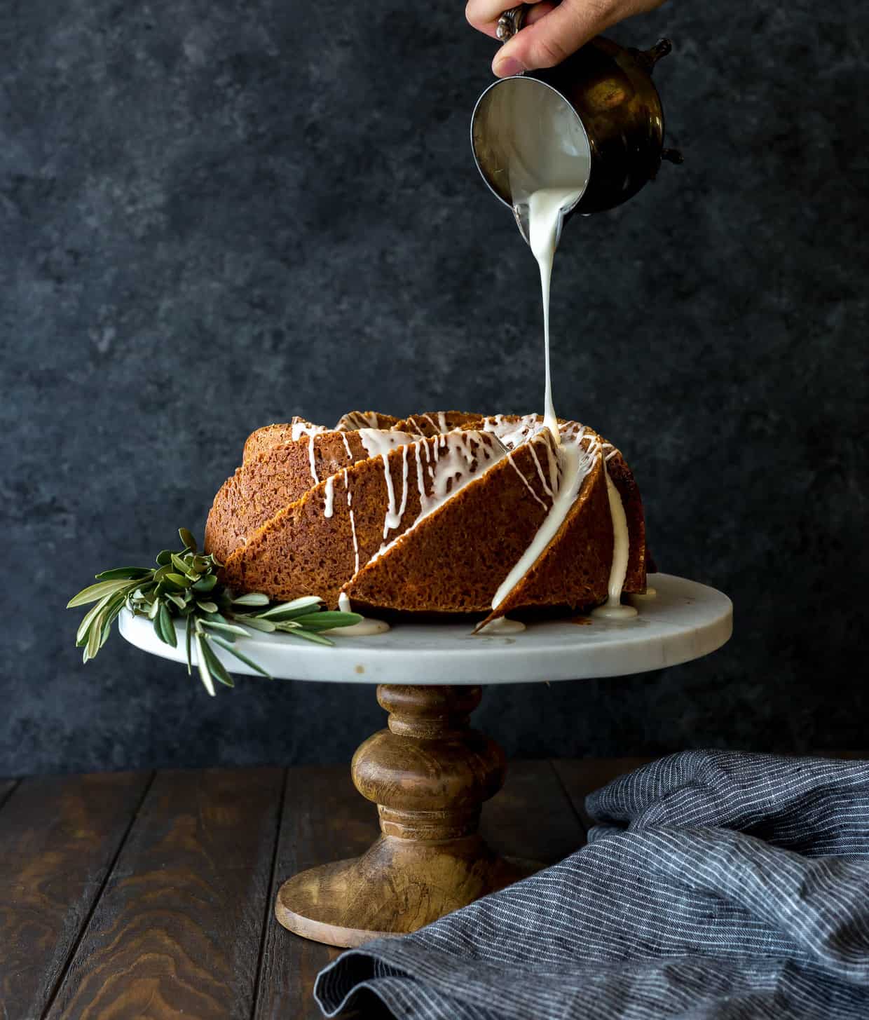 A beautiful gingerbread bundt cake on a cake stand, with olive branches laying at the base of the cake.