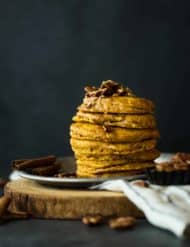 A stack of 6 pumpkin pancakes sitting on a metal tin, with cinnamon brown butter pecan syrup drizzling down the sides.