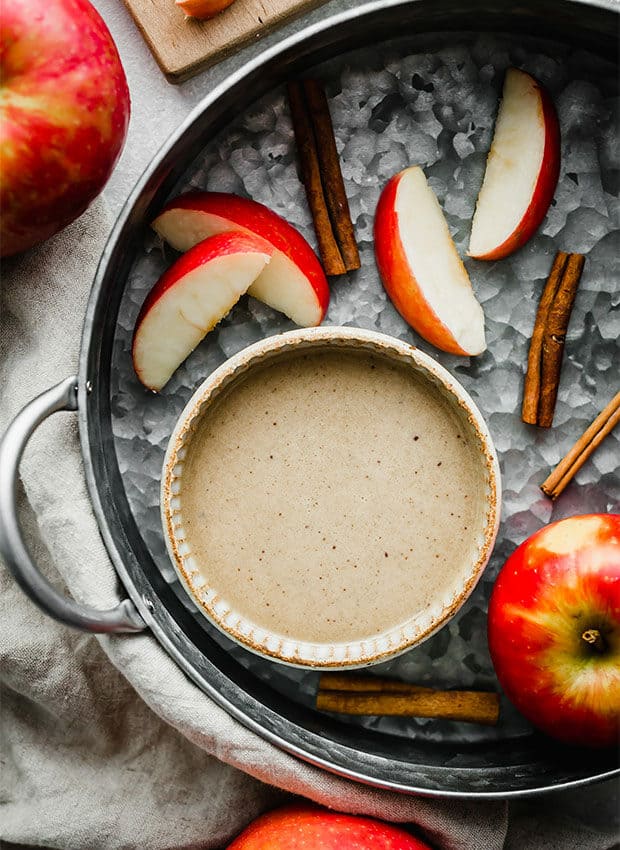 A bowl of caramel apple dip surrounded by cinnamon sticks and sliced apples.