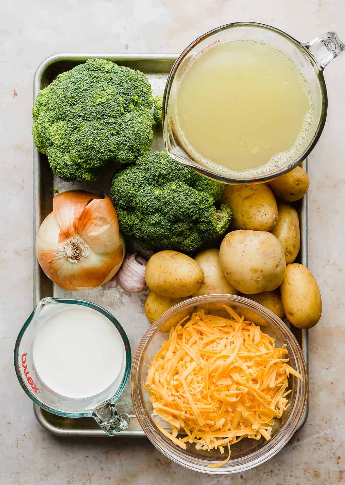 A baking sheet full of ingredients used to make Broccoli Potato Soup: cream, cheese, potatoes, chicken broth, broccoli.