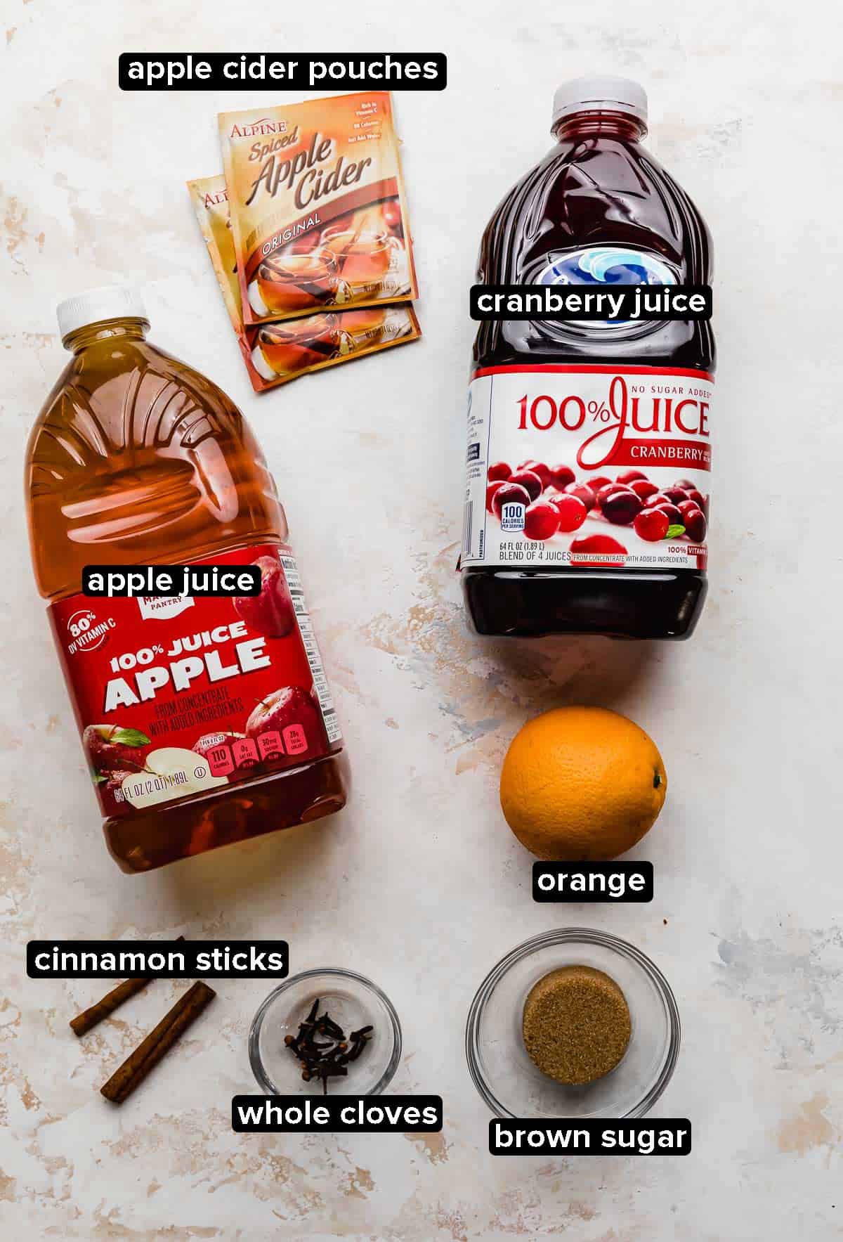 Cranberry Apple Cider ingredients on a white textured background: brown sugar, orange, cranberry juice, apple juice, spiced cider, cloves, and cinnamon sticks.