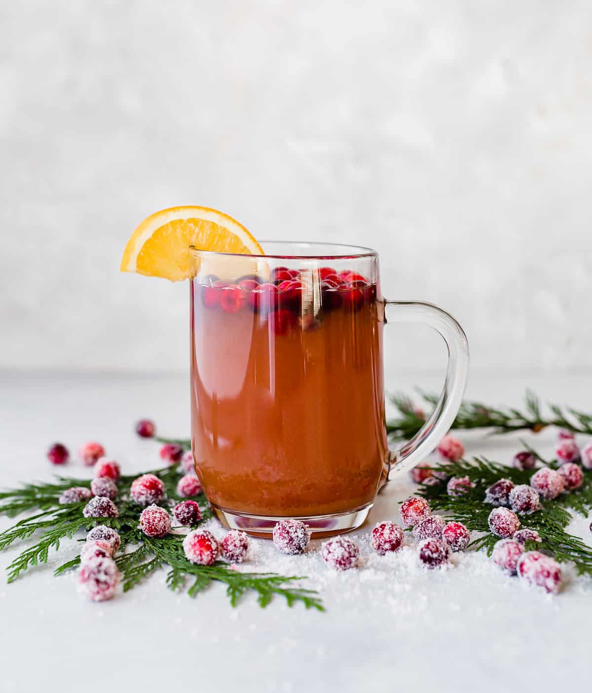 A glass mug filled with Cranberry Apple Cider against a white background with sugared cranberries on the table near the mug.