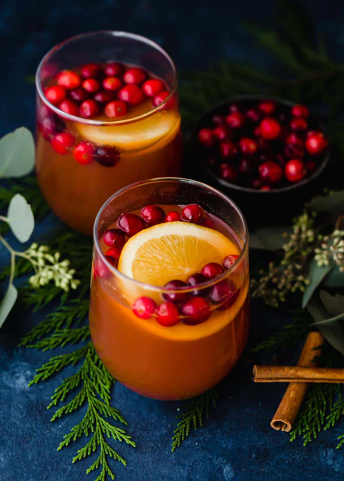 Two cups of Cranberry Apple Cider filled with fresh cranberries and orange slice on a dark blue background.
