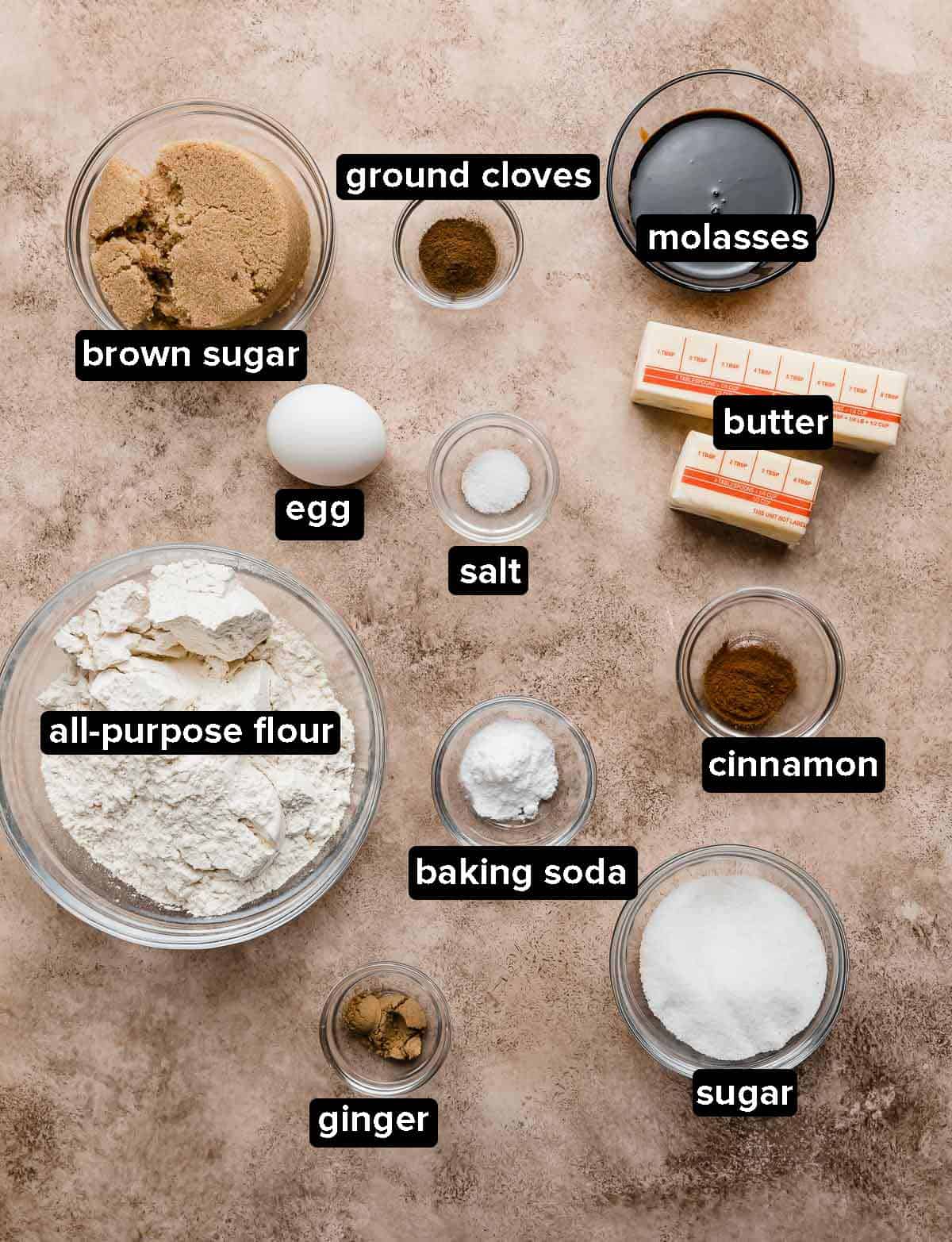 Gingersnap cookie ingredients portioned into glass bowls on a light brown textured background.