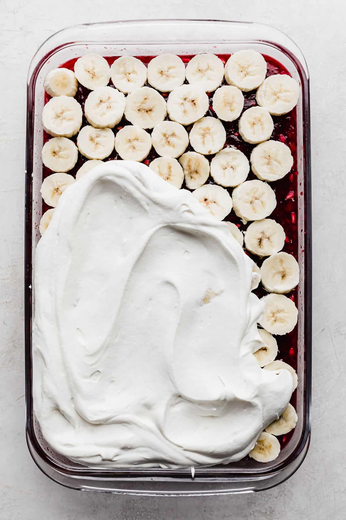 Whipped cream spread overtop half of the sliced bananas that are overtop the red Pomegranate Jell-O.