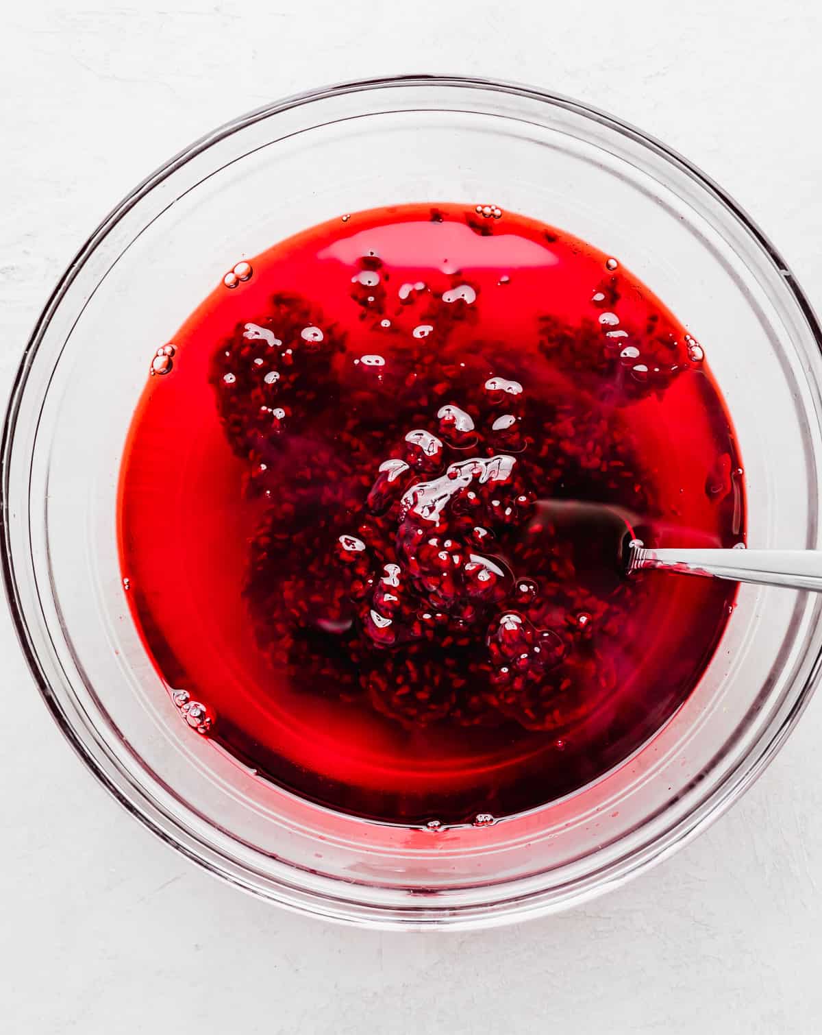 Bright red liquid jell-o with a raspberry topping sauce being stirred into it.