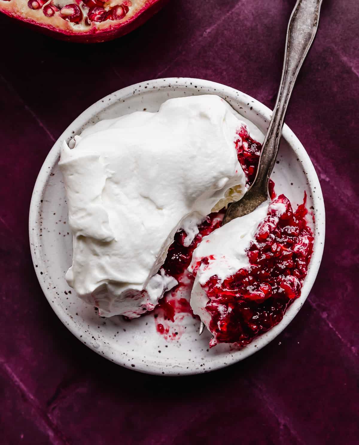 A slice of whipped cream topped Pomegranate Jell-O salad on a white plate on a deep maroon colored background.