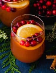 A glass cup with Cranberry Apple Cider, fresh cranberries, and an orange slice in it.