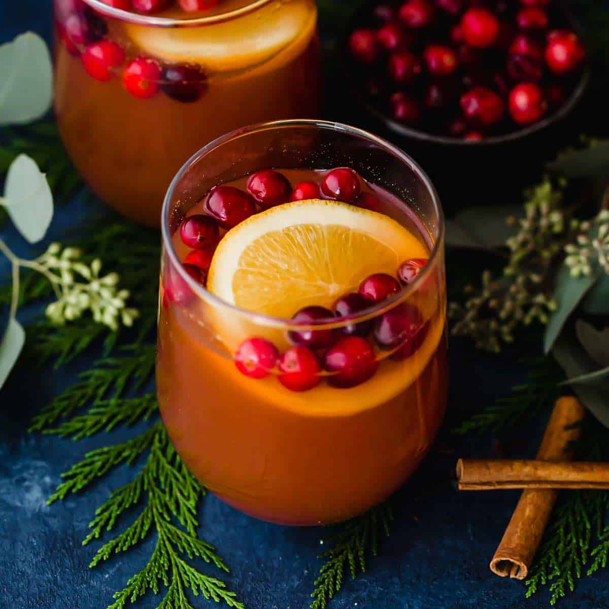 A glass cup with Cranberry Apple Cider, fresh cranberries, and an orange slice in it.