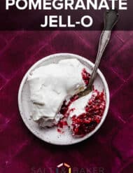 Whipped topping covered Christmas Pomegranate Jell-O on a white plate that's sitting on a plum colored background.