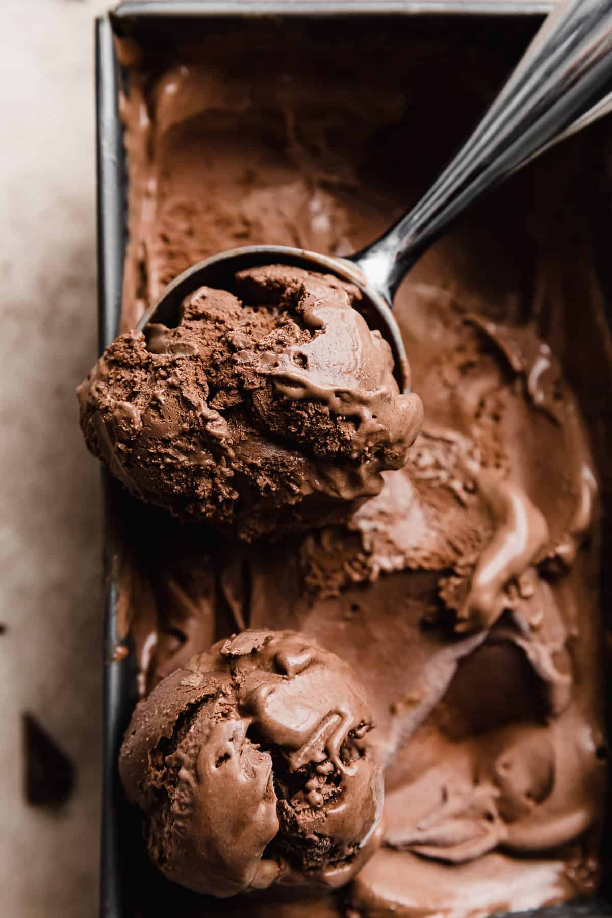 An ice cream scooper scooping out dark chocolate ice cream from a rectangle container.