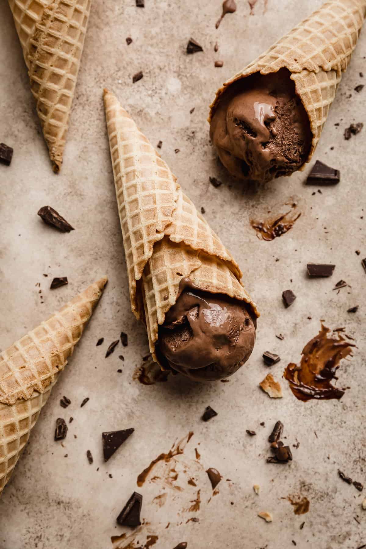 Homemade Chocolate Ice Cream in a waffle cone laying flat on a light brown textured background with chopped chocolate scattered around.