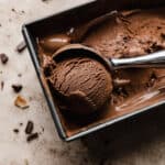The best chocolate ice cream in a bread pan with an ice cream scooper scooping out a ball of the brown ice cream.