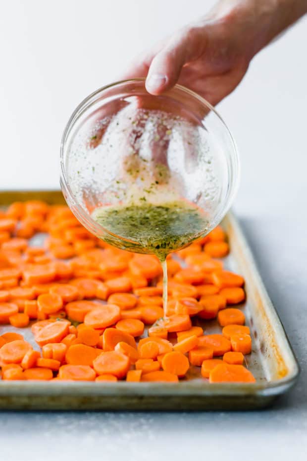 Melted butter, garlic, parsley, and parmesan being drizzled over a baking sheet covered in chopped carrots.