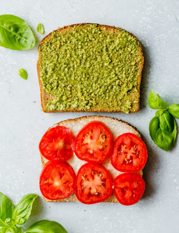 Two slices of bread, one covered in basil pesto and the other has 6 slices of fresh Roma tomatoes on top.