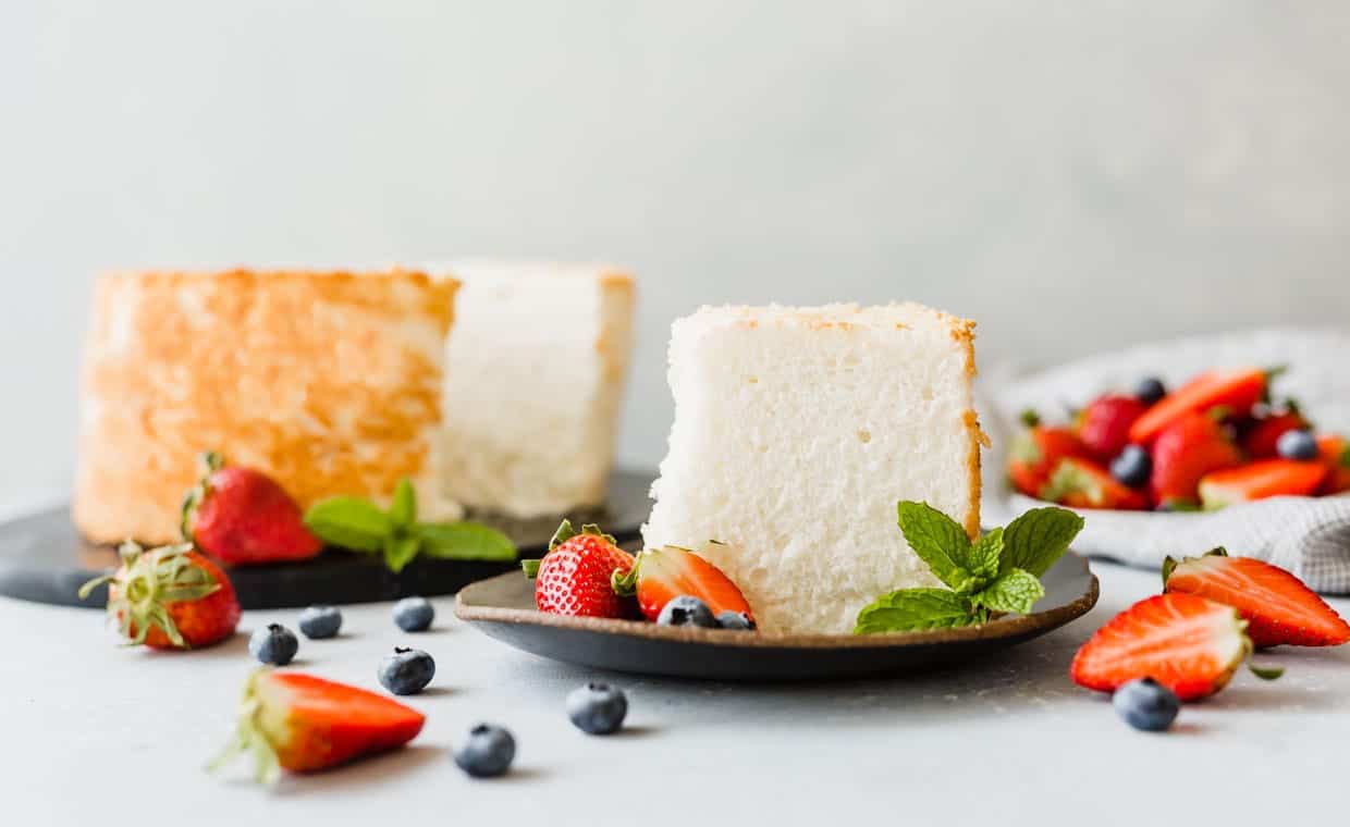 A slice of angel food cake on a plate, with strawberries and blueberries throughout the scene.