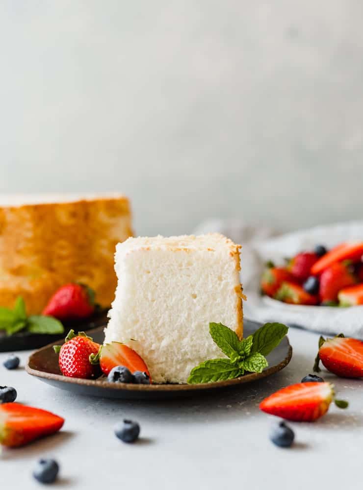 A slice of angel food cake on a black plate, with strawberries and blueberries scattered throughout the scene.