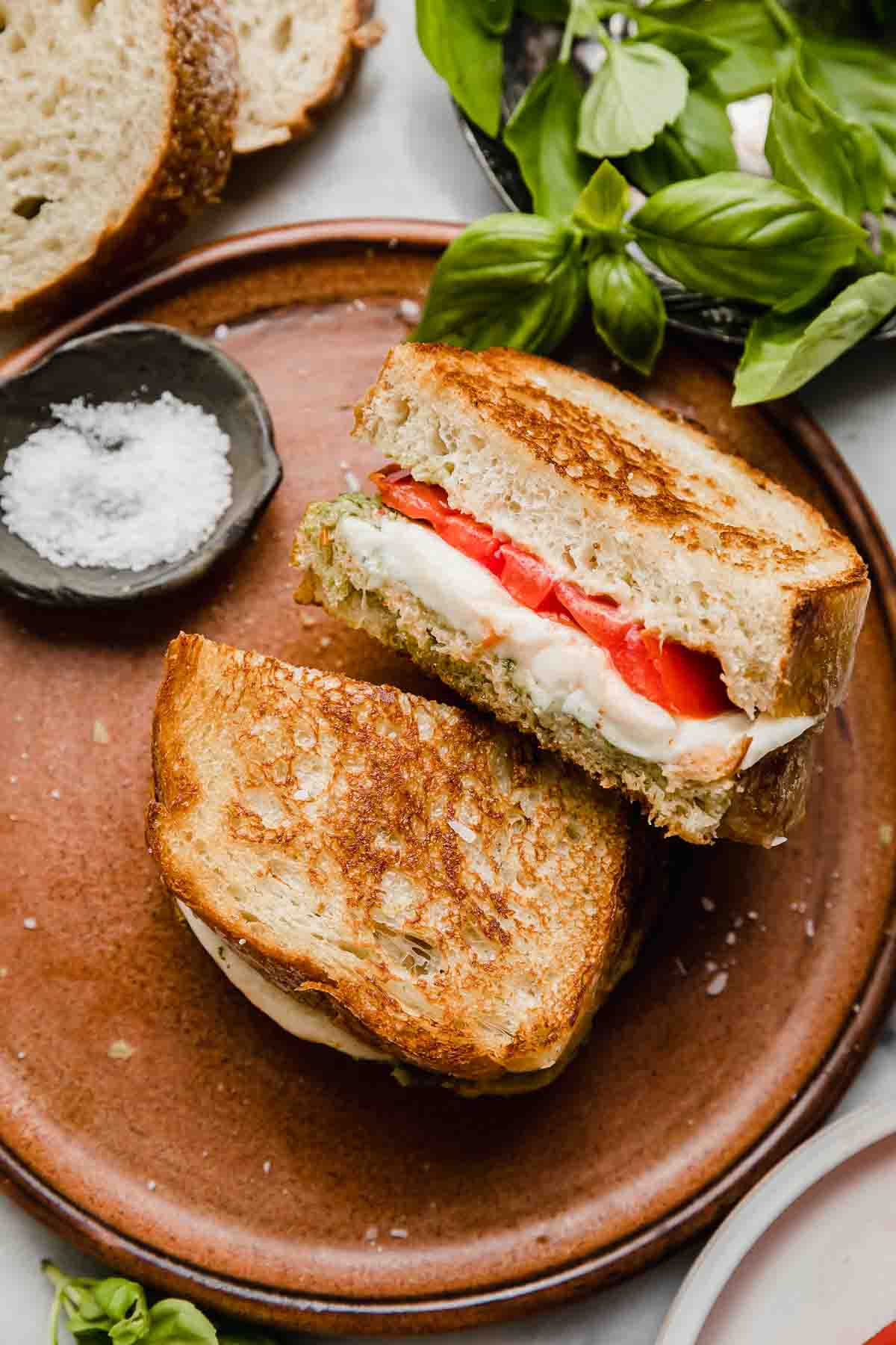 A Pesto Mozzarella Sandwich with tomato, sliced in half on a brown plate.