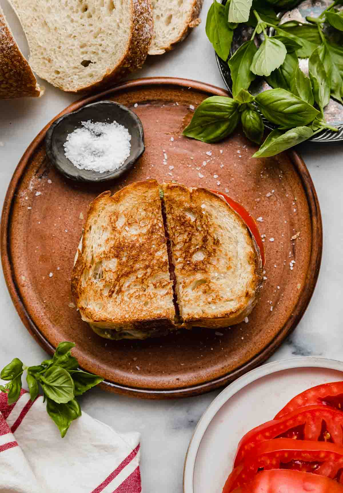 Pesto Mozzarella Sandwich on a brown plate, sliced in half with fresh basil in a bowl at the right corner of the photo.