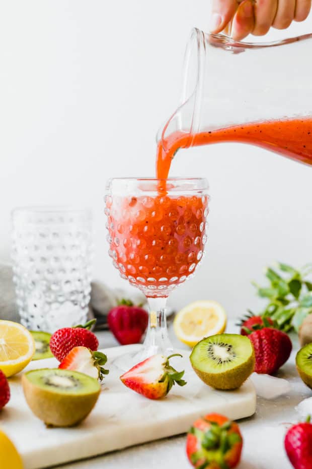 A pitcher of Strawberry Kiwi Lemonade being poured into a clear glass with ice in it.