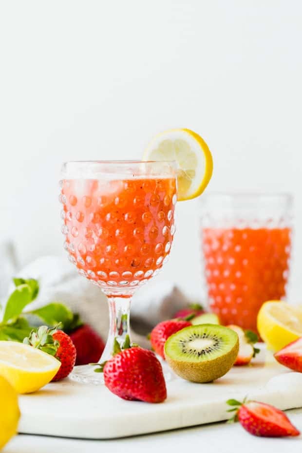 Two clear glasses full of strawberry kiwi lemonade with fresh strawberries, kiwis, and lemons surrounding the glasses.