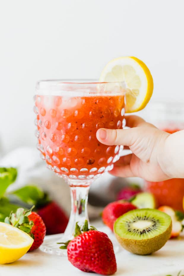 A clear glass full of strawberry kiwi lemonade, and a young boys hand grabbing the glass. 