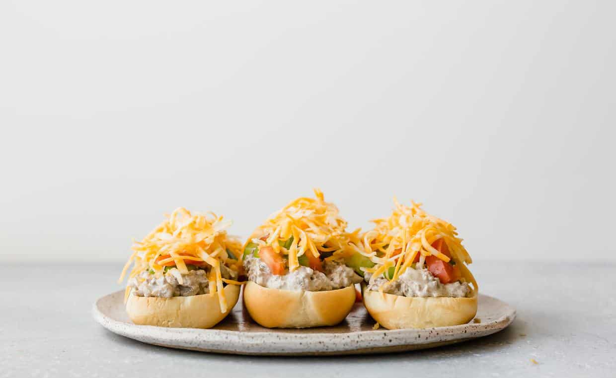Three hoagie buns on a plate, each topped with beef stroganoff, diced bell peppers, tomatoes, and shredded cheese.