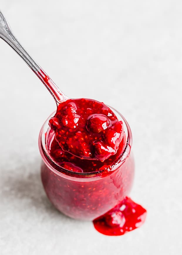 A glass jar on a white background, overflowing with raspberry sauce.