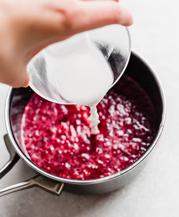 A cornstarch slurry being poured into a saucepan full of a raspberry compote.