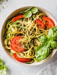 Zucchetti Pasta in a white bowl with nutritional yeast, tomatoes, and fresh basil.