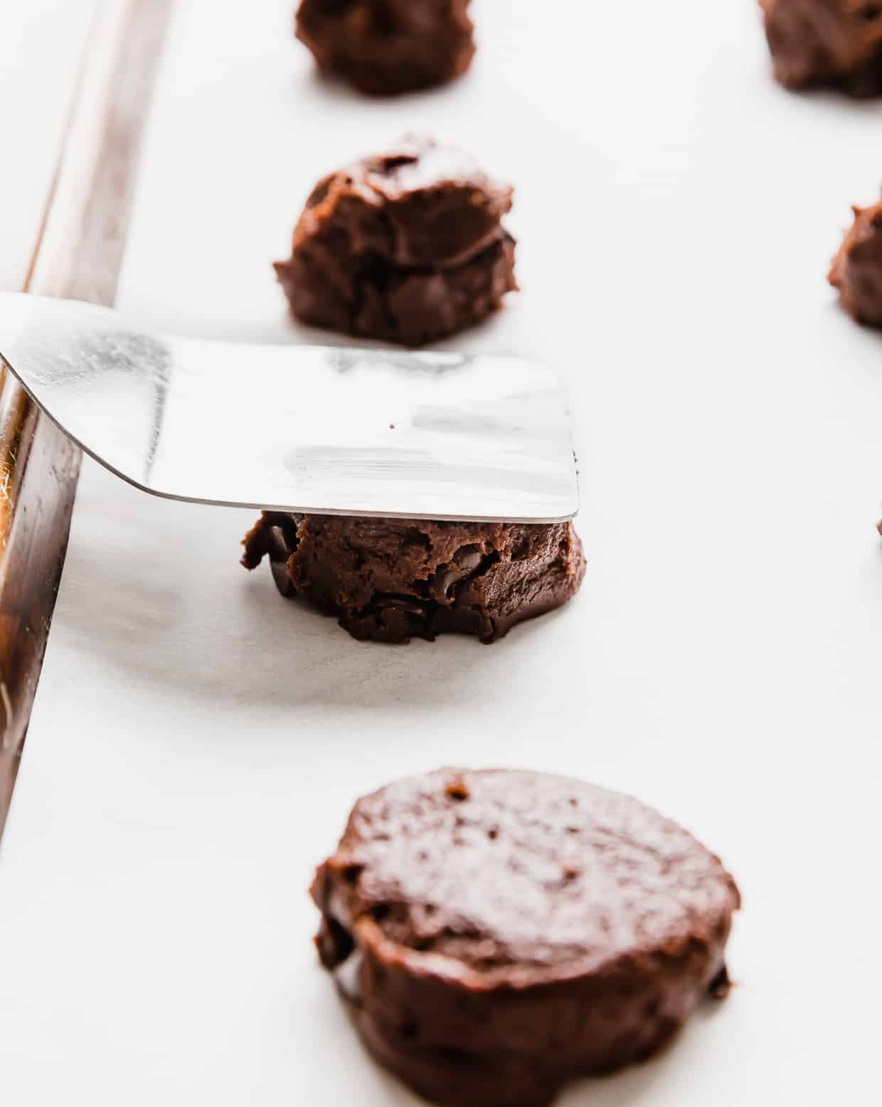 A metal spatula pressing down on a chocolate cookie dough ball on a baking sheet.
