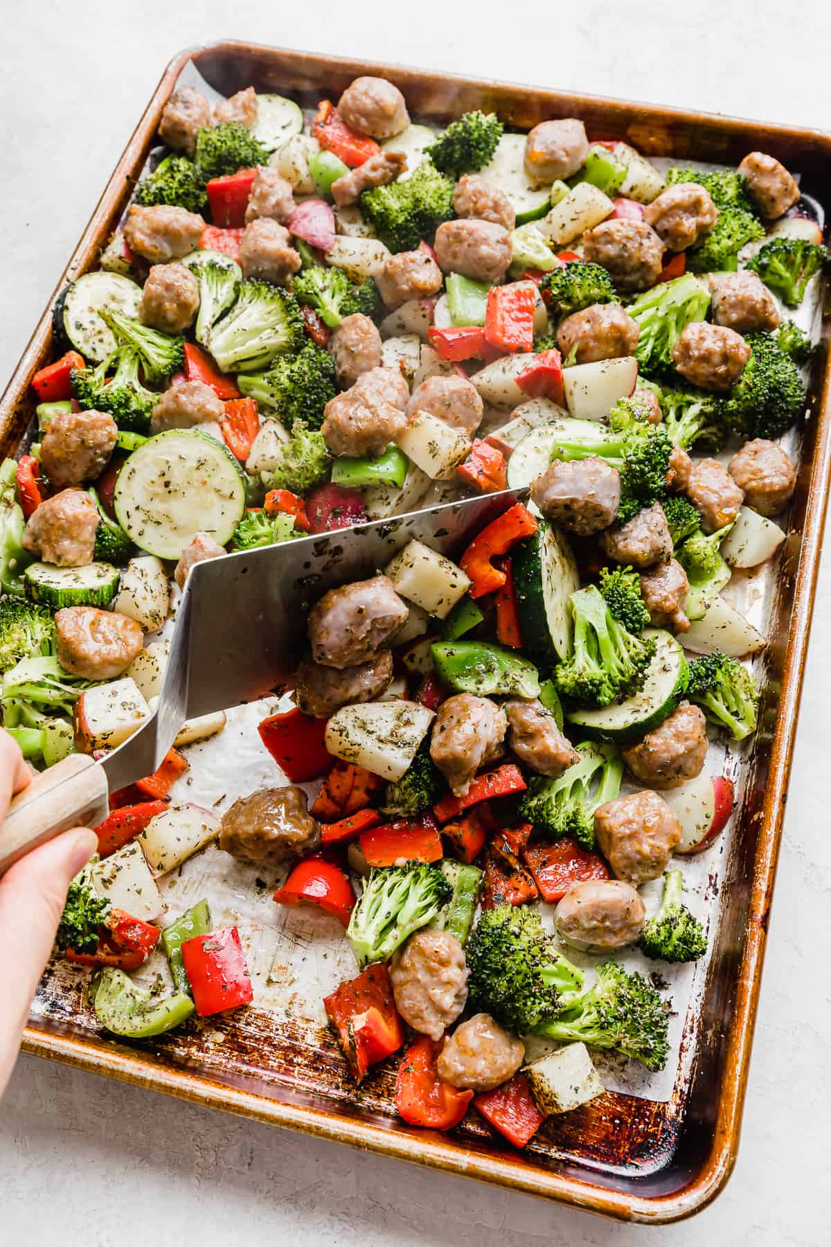 A metal spatula turning over sausage and veggies on a sheet pan.
