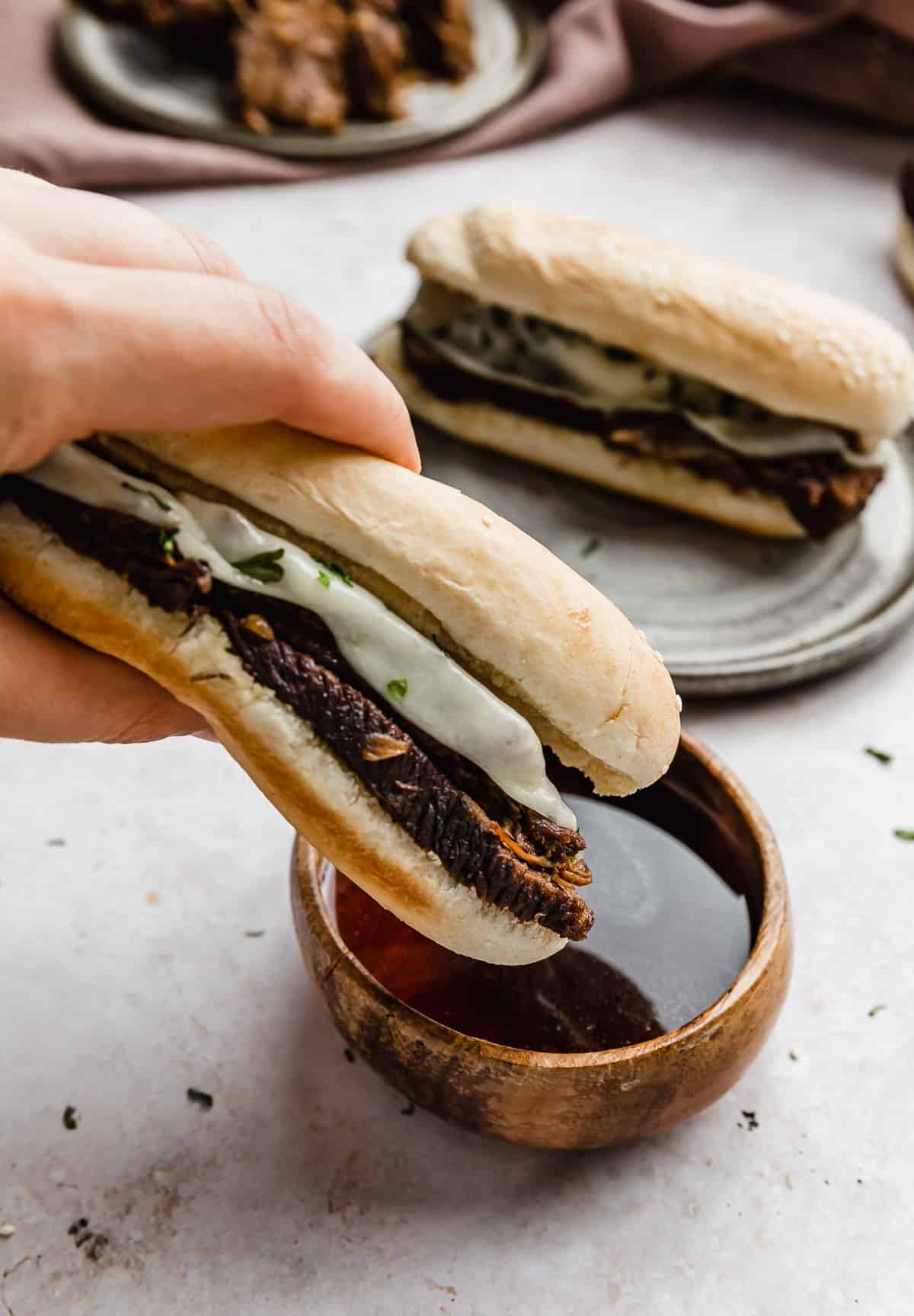 A tender French dip sandwich topped with provolone cheese being dipped into a brown bowl full of au juice.