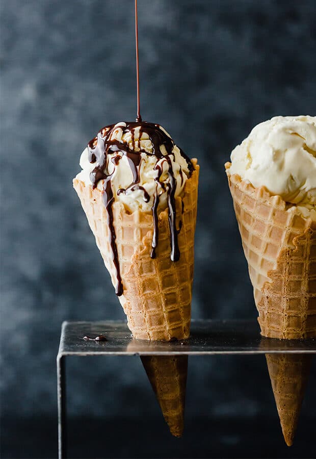 An waffle cone full of homemade vanilla ice cream, with chocolate sauce being drizzled over the ice cream.