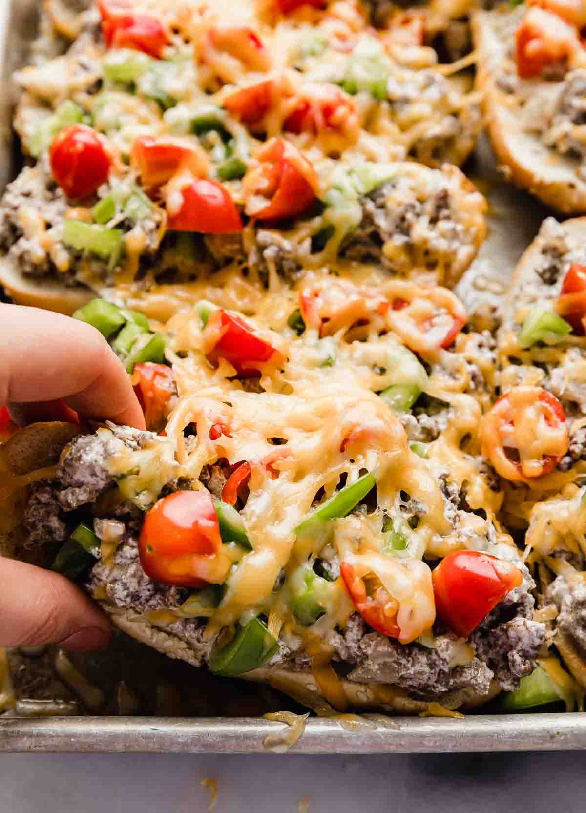 A hand grabbing a Beef Stroganoff Sandwich that's topped with ground beef, tomatoes, peppers, and shredded melted cheese.