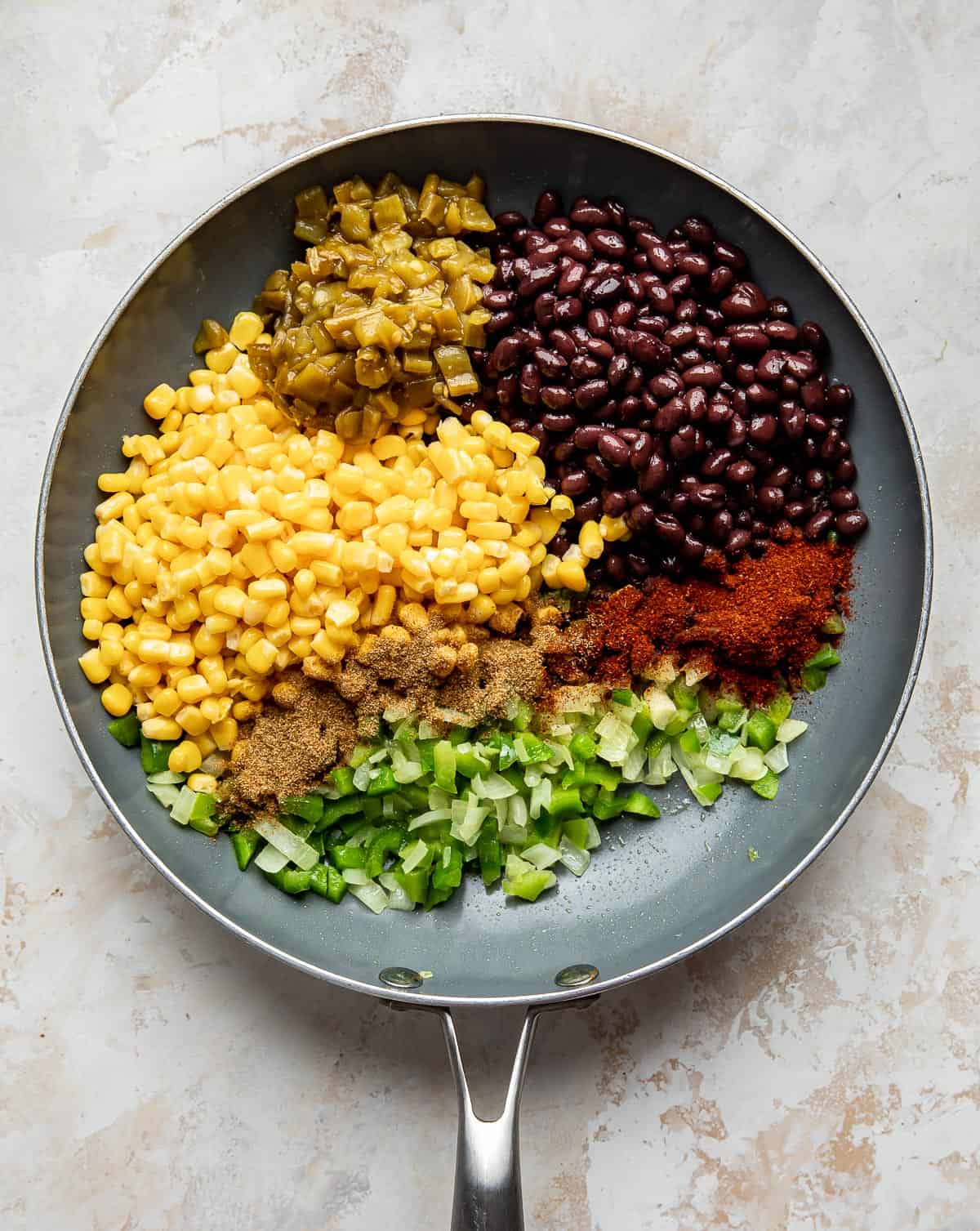A gray skillet with corn, green pepper, black beans, chili powder, and diced green chilies in it.