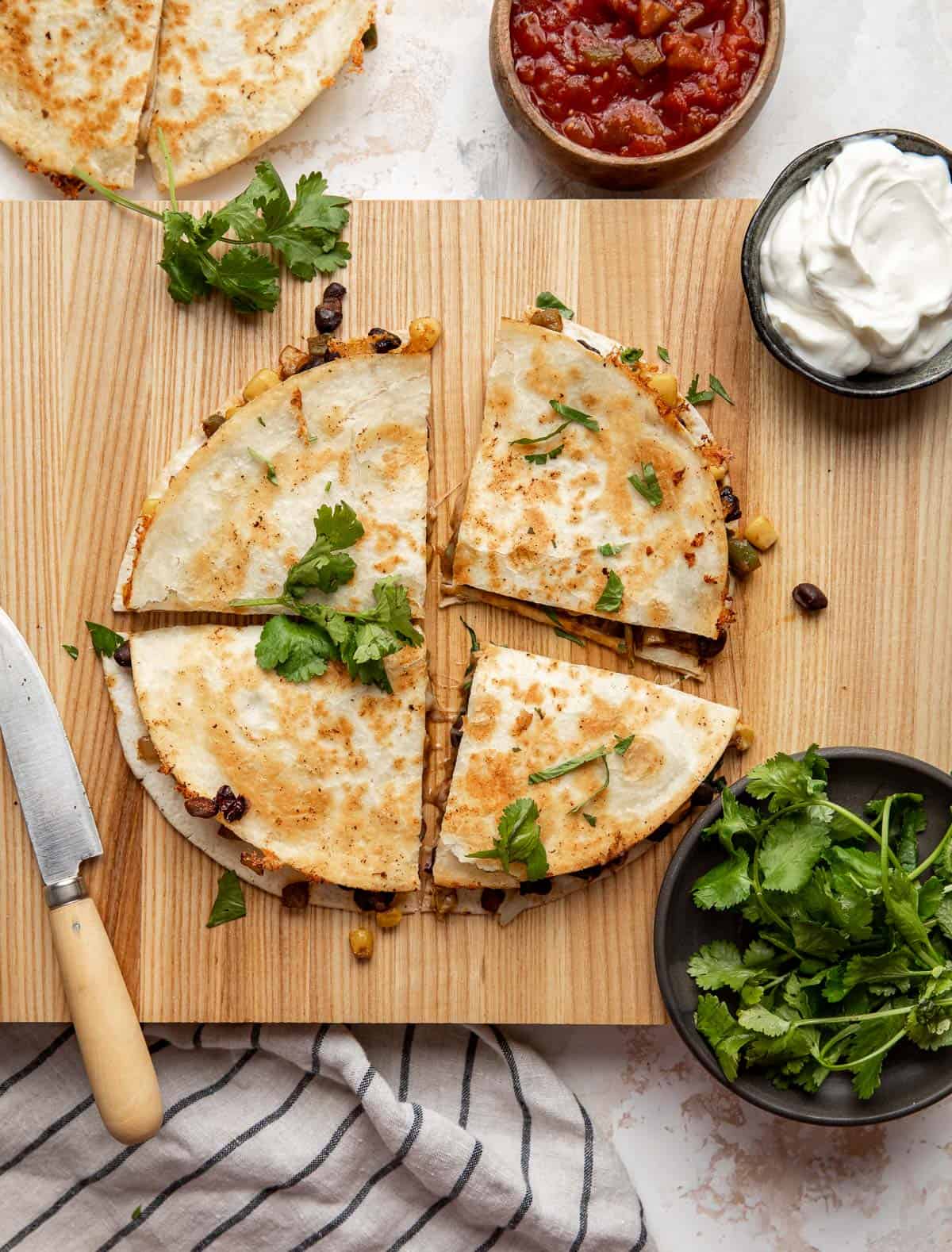 Black Bean and Corn Quesadilla on a wooden cutting board surrounded by sour cream, cilantro, and salsa. 